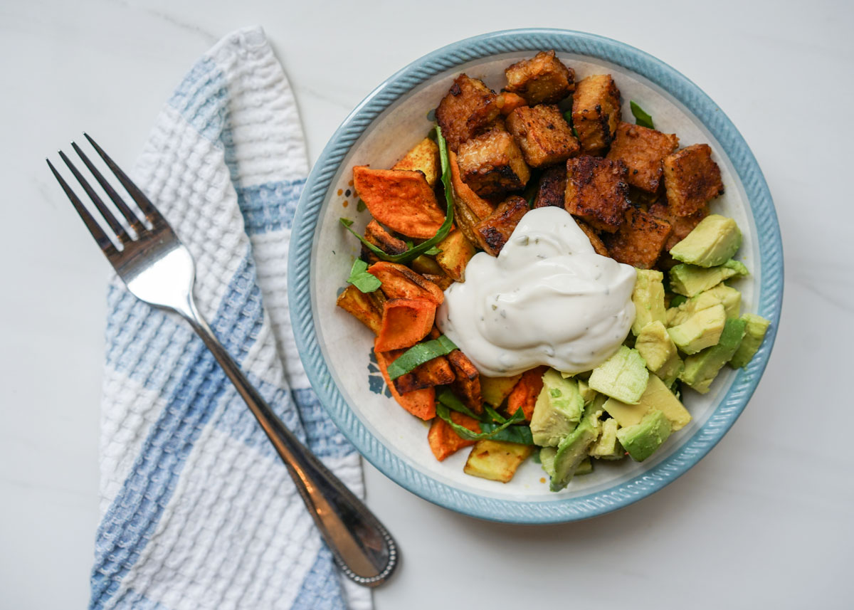 Tempeh and roasted root vegetable buddha bowl in a bowl on white marble counter.